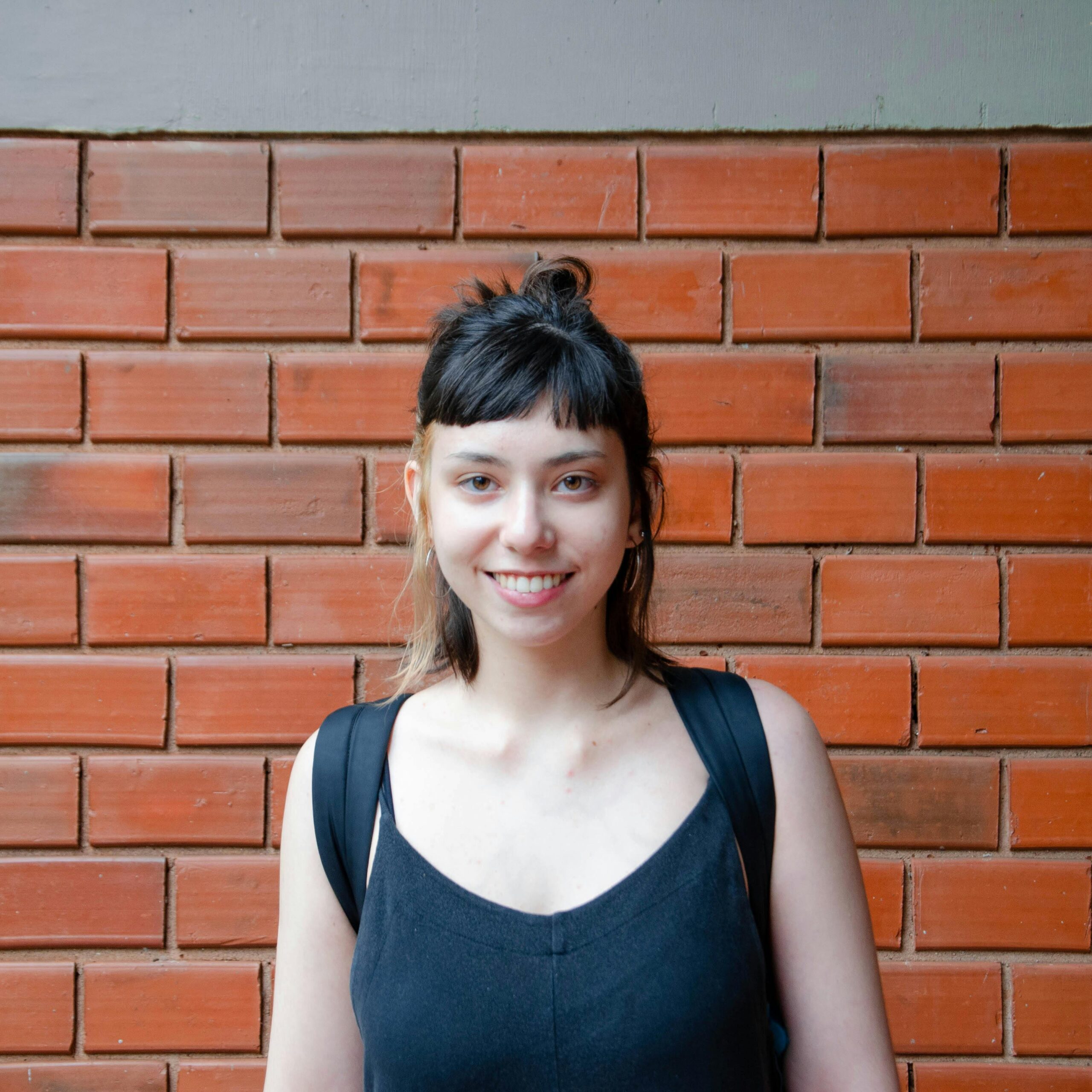 Portrait of a young woman with a backpack smiling against a brick wall. Casual and relaxed.