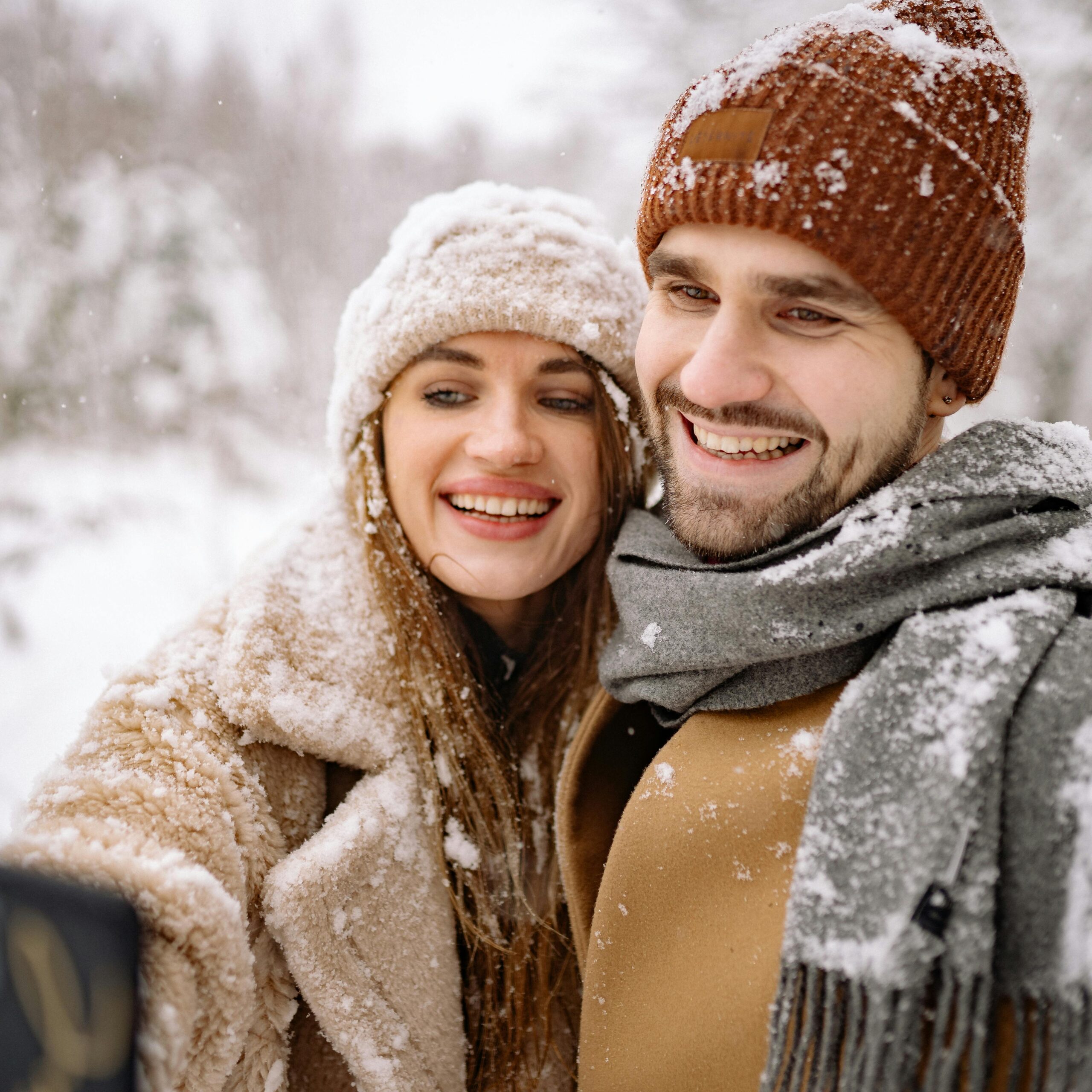 Happy couple in winter clothing, taking a selfie in a snowy outdoor setting.